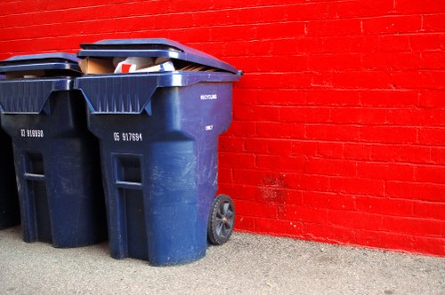Two large dark blue plastic wheelie bins with black lids are positioned on a paved surface next to a red brick wall. The bins are side by side, with the left bin slightly in front of the right and appear to contain mixed waste, with some paper and plastic visible above the lids. The texture of the bins is smooth and slightly glossy, reflecting light. The black lids are securely closed, and the bins are equipped with black wheels at the rear for mobility. The environment suggests an outdoor area, possibly a back alley or service space associated with a commercial property in Chessington. The background features a vibrant red brick wall, and the scene is evenly lit without visible shadows or weather effects. This setup aligns with typical arrangements used for rubbish collection and waste removal services, such as those provided by Commercial Waste Chessington, focusing on general waste or rubbish management for local businesses or residential properties in the area.