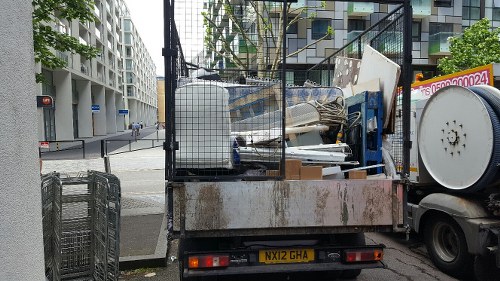 A flatbed truck parked on a city street in Chessington, loaded with a mix of large, bulky rubbish items including white and beige panels, metal and plastic furniture parts, and assorted debris, with rusted areas on the truck’s metal surface. A metal fence surrounds the load, with some items leaning against it. To the right, a large white spool or drum is visible, partially attached to the truck, while the background features modern residential and commercial buildings with glass balconies and windows, and an intersection with traffic lights. The pavement and street are visible, along with a glimpse of the corner of a nearby building on the left. The scene suggests a waste collection or rubbish removal activity by Commercial Waste Chessington, situated in a busy urban area within or near the post code area associated with Chessington, providing a clear view of rubbish awaiting disposal.