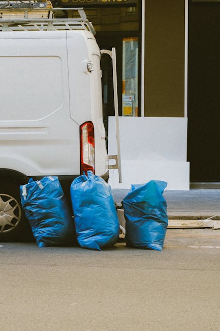 A white commercial van parked on a paved street next to a sidewalk, with three large blue rubbish bags placed on the ground against the vehicle's rear, filled with waste materials. The van features a 
