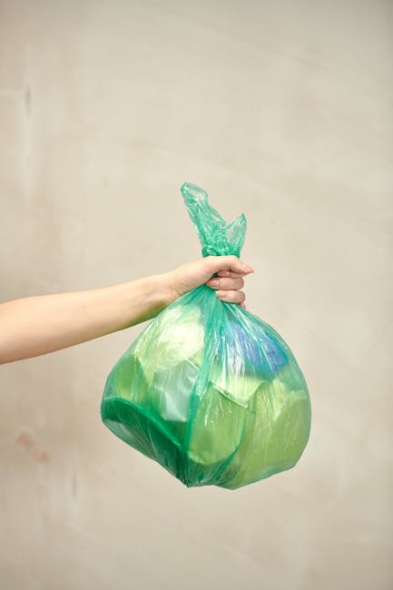 A white commercial van parked on a paved street next to a sidewalk, with three large blue rubbish bags placed on the ground against the vehicle's rear, filled with waste materials. The van features a 
