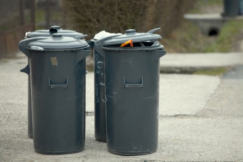 Two large, dark grey plastic wheelie bins with closed lids are positioned on a residential driveway or pavement surface, situated side by side in front of a blurred background featuring some greenery and a concrete pathway. The left bin has a smooth, matte finish with a slightly raised handle on the front, while the right bin's lid is partially open, revealing a small orange item and some other waste partially visible inside. The bins are used for waste collection in the local area, and their placement suggests routine rubbish disposal. The environment appears to be a suburban setting, consistent with typical residential waste management practices, and the bins are branded with minimal markings, characteristic of standard refuse containers used by commercial waste services such as Commercial Waste Chessington. The lighting indicates daytime with natural illumination.
