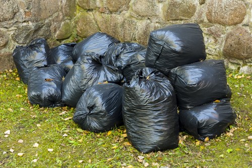 A collection of eleven large black plastic rubbish sacks filled with waste, piled on a patch of grass near a stone wall. The sacks are tightly tied at the top and have a shiny, crinkled texture. The background features irregularly shaped, weathered stone blocks forming the wall, indicating an outdoor setting. The surrounding ground is covered with small fallen leaves and bits of foliage, characteristic of an urban or suburban environment. The image conveys a scene of waste prepared for rubbish collection, consistent with commercial waste management services in Chessington. The setting suggests an outdoor area adjacent to a property or business premises, with natural light illuminating the scene. The arrangement and appearance of the sacks reflect typical rubbish removal practices, emphasizing the importance of proper waste disposal managed by companies like Commercial Waste Chessington.