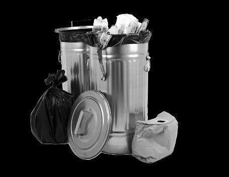 A large cylindrical metal rubbish bin with a domed lid placed in an indoor or outdoor environment, surrounded by black background. The bin is lined with a black plastic bag that is filled with assorted waste materials, including crumpled paper and plastic items, which protrude from the top. To the left of the bin is a tied black garbage bag, leaning against its side, while on the right side, a partially crumpled white paper or plastic bag is positioned on the ground. The bin lid, resting against the base, is metallic with a smooth, slightly reflective surface. The scene conveys a professional waste management setup, commonly associated with commercial rubbish removal services, such as those offered by Commercial Waste Chessington, and emphasizes proper waste containment and disposal practices in a typical setting near a business or property in Chessington, Surrey.